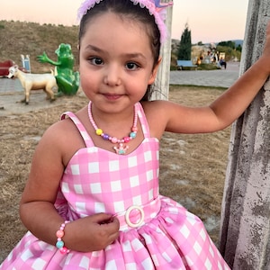 May include: A young girl wearing a pink and white gingham dress with a white belt and a pink crown headband. She is also wearing a colorful beaded necklace and a bracelet.