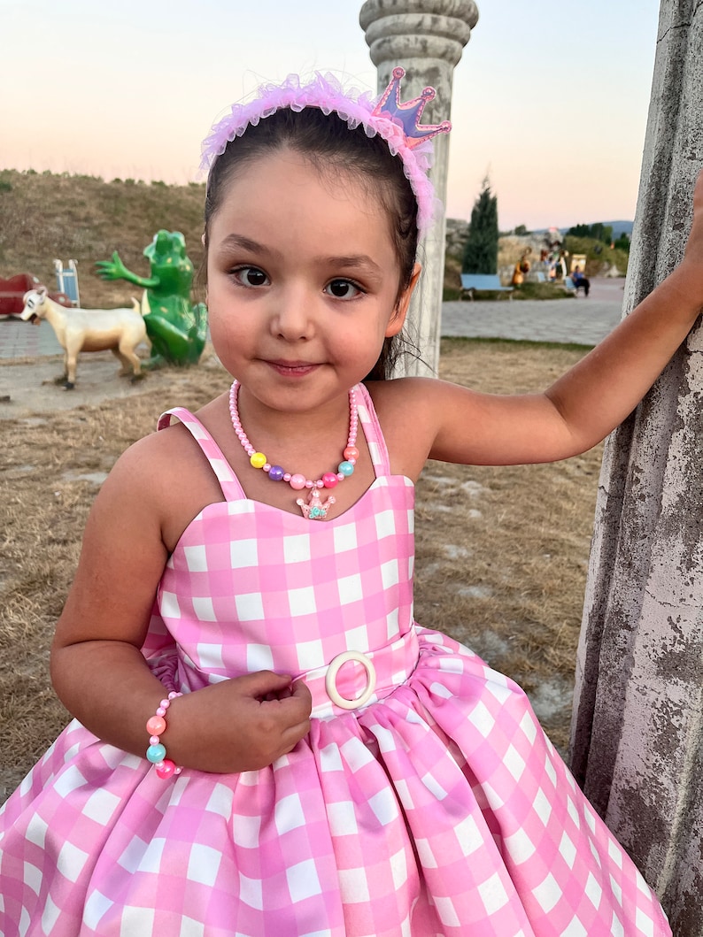 May include: A young girl wearing a pink and white gingham dress with a white belt and a pink crown headband. She is also wearing a colorful beaded necklace and a bracelet.
