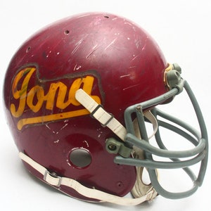 K&ouml;nnte beinhalten: Ein maroonfarbener Footballhelm mit einem wei&szlig;en und gelben "Ford"-Logo. Der Helm hat ein graues Gesichtsgitter.