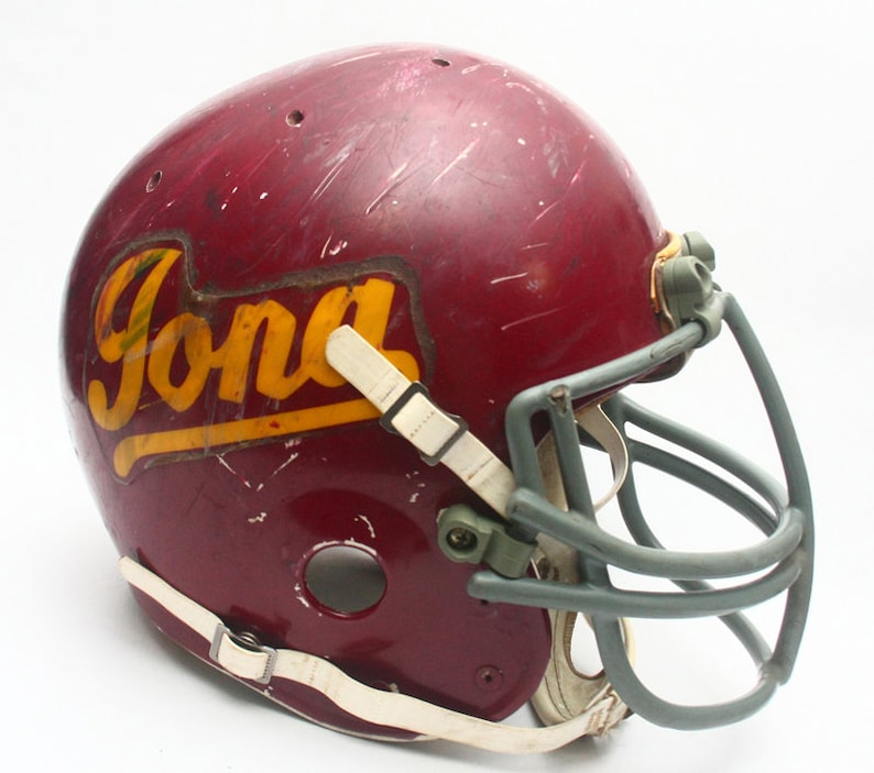 K&ouml;nnte beinhalten: Ein maroonfarbener Footballhelm mit einem wei&szlig;en und gelben "Ford"-Logo. Der Helm hat ein graues Gesichtsgitter.