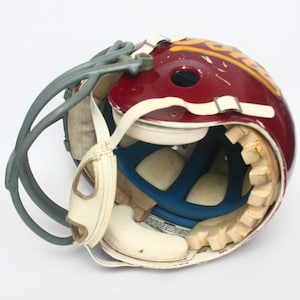 K&ouml;nnte beinhalten: Ein burgunderfarbener Footballhelm mit einem wei&szlig;en und goldenen Logo. Der Helm ist ge&ouml;ffnet und zeigt die Innenpolsterung und ein blaues Futter.