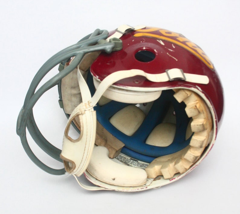 K&ouml;nnte beinhalten: Ein burgunderfarbener Footballhelm mit einem wei&szlig;en und goldenen Logo. Der Helm ist ge&ouml;ffnet und zeigt die Innenpolsterung und ein blaues Futter.