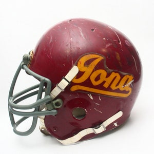 K&ouml;nnte beinhalten: Ein maroonfarbener Footballhelm mit einem grauen Gesichtsgitter und einem goldenen "Iona"-Logo.