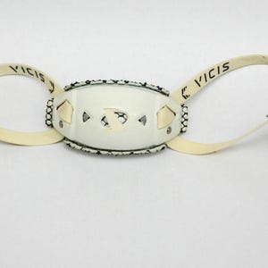 Pode incluir: Máscara de olhos em couro creme com a palavra "VICIS" impressa em cada tira. A máscara apresenta uma secção central oval com recortes geométricos e uma borda decorativa. Ferragens e tiras de metal completam o design.