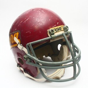 K&ouml;nnte beinhalten: Ein burgunderfarbener Footballhelm mit einem wei&szlig;en Gesichtsgitter und einem gelben und goldenen "GINA"-Logo an der Seite. Der Helm hat ein graues Gesichtsgitter mit einem "BIKE"-Aufkleber oben.