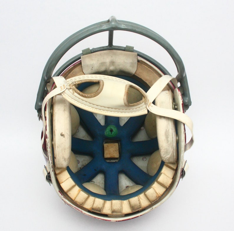 K&ouml;nnte beinhalten: Ein Vintage-Footballhelm mit rot-wei&szlig; gestreiftem &Auml;u&szlig;eren. Der Helm hat ein graues Metallgesichtsschutzgitter und ein wei&szlig;es Lederinnere mit Polsterung.