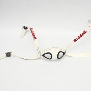 Puede incluir: Una correa de barbilla blanca para casco de fútbol Riddell con orificios negros para los ojos y el logotipo de Riddell en rojo. La correa tiene cierres metálicos y está diseñada para asegurar un casco de fútbol.