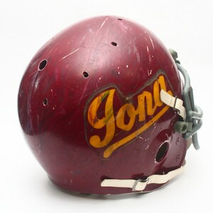 K&ouml;nnte beinhalten: Ein maroonfarbener Footballhelm mit einem wei&szlig;en Gesichtsgitter und einem gelben und goldenen "Jona"-Logo an der Seite.