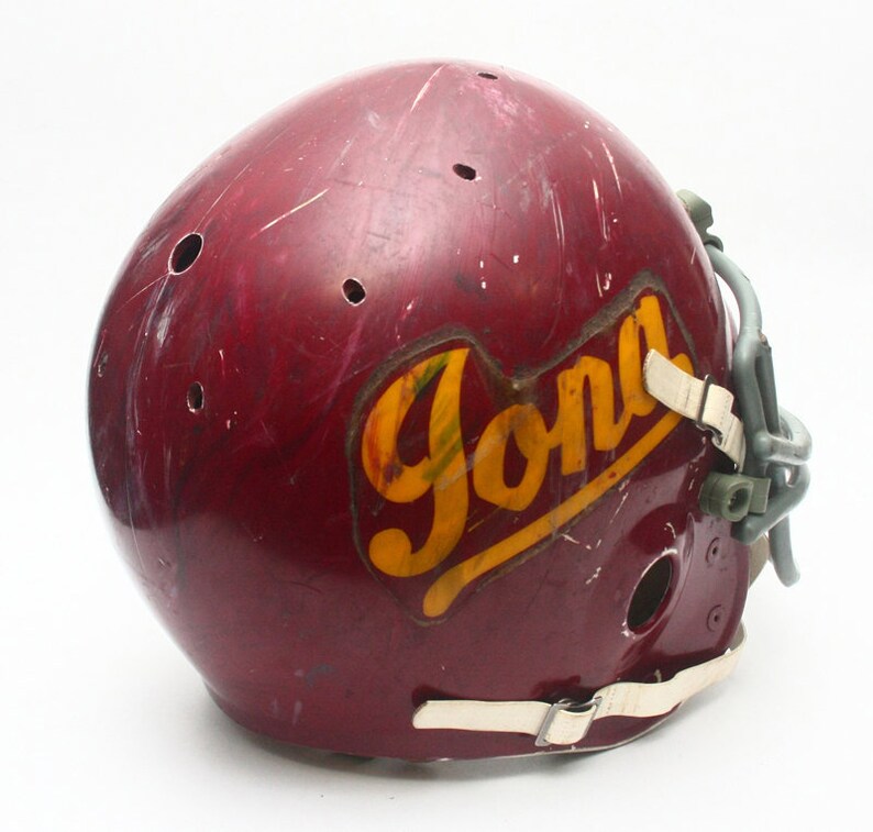 K&ouml;nnte beinhalten: Ein maroonfarbener Footballhelm mit einem wei&szlig;en Gesichtsgitter und einem gelben und goldenen "Jona"-Logo an der Seite.