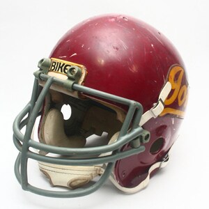 K&ouml;nnte beinhalten: Ein burgunderfarbener Footballhelm mit einem grauen Gesichtsgitter und einem wei&szlig;en und goldenen "JOKER"-Logo an der Seite. Der Helm hat einen "BIKE"-Aufkleber auf der Vorderseite.