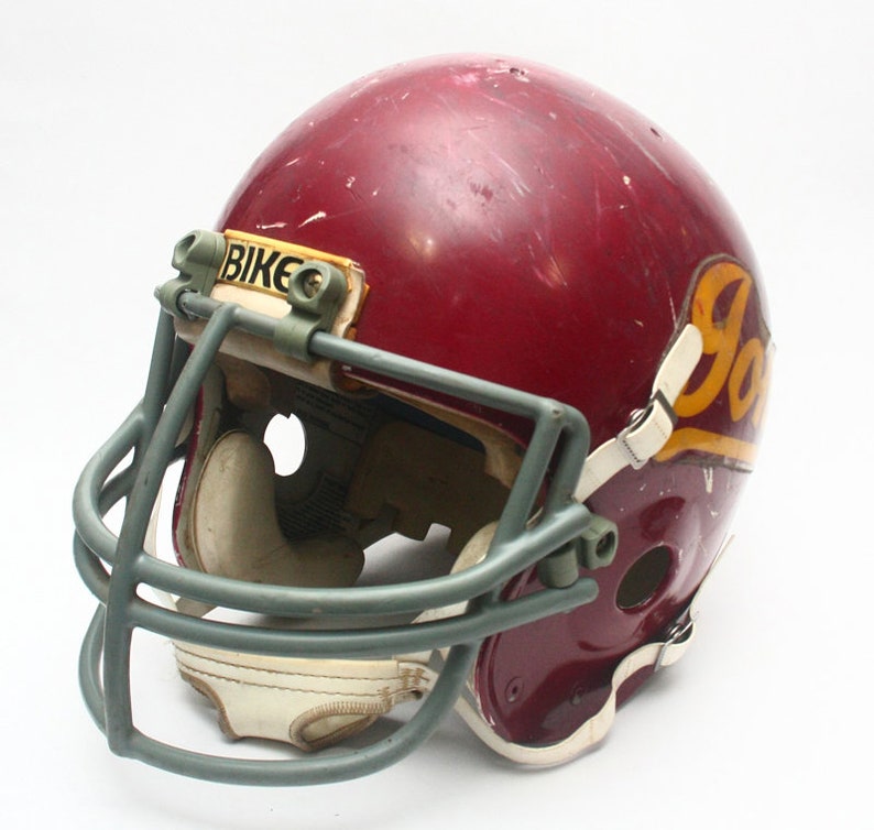 K&ouml;nnte beinhalten: Ein burgunderfarbener Footballhelm mit einem grauen Gesichtsgitter und einem wei&szlig;en und goldenen "JOKER"-Logo an der Seite. Der Helm hat einen "BIKE"-Aufkleber auf der Vorderseite.