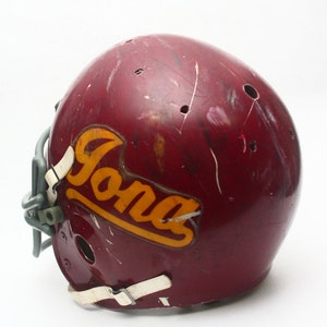 K&ouml;nnte beinhalten: Ein maroonfarbener Footballhelm mit einem wei&szlig;en Gesichtsgitter und einem gelben und goldenen "Iona"-Aufkleber an der Seite.
