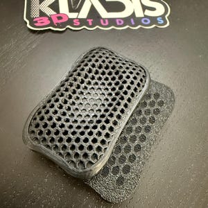 May include: Two black 3D-printed rectangular objects with a honeycomb pattern. One is stacked on top of the other. The objects are on a dark surface. A sticker with the text "KLADIS 3D STUDIOS" is in the background.