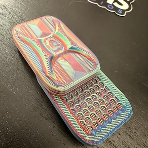 Op de afbeelding: Een veelkleurig, 3D-geprint klaptelefoonspeelgoed. De telefoon is voornamelijk blauw, rood en groen, met een textuuroppervlak en een honingraatpatroon op de onderste helft. De bovenste helft heeft een uniek ontwerp.