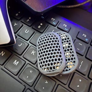 Könnte beinhalten: Zwei graue, perforierte, rechteckige Objekte mit abgerundeten Kanten liegen auf einer schwarzen Computertastatur. Die Objekte haben ein gitterartiges Muster aus kleinen Löchern. Die Tastatur ist im Fokus, mit Tasten wie 'Alt', 'K' und 'M' sichtbar.