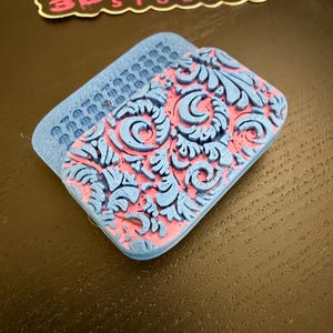 May include: A blue and pink decorative item with an ornate floral design. The item has a rectangular shape with a textured surface and the text "KLADIS 3D STUDIOS" in the upper left corner.
