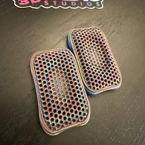 May include: Two rectangular, iridescent 3D-printed objects with a honeycomb pattern. The objects have a blue border and are placed on a dark surface. A sticker with the text "KLADIS 3D STUDIOS" is in the background.