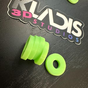 May include: Bright green 3D-printed objects, including a cylindrical piece with stacked rings and two circular pieces with holes. The objects are on a dark surface, with a sticker that reads "KLADIS 3D STUDIOS".