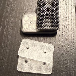 May include: A rectangular black object with a textured honeycomb pattern on its top surface, paired with two clear, rectangular plastic plates. Each clear plate features two circular holes, and the black object shows a visible central screw.