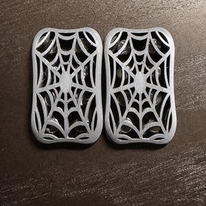 May include: Two silver-colored, rectangular decorative pieces with a spiderweb design. The spiderweb pattern is cut out, revealing a dark interior. The pieces are placed side by side on a dark brown surface.