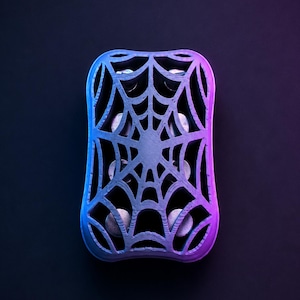 Könnte beinhalten: Ein rechteckiger, silberfarbener Gegenstand mit einem Spinnennetz-Design. Das Objekt hat einen blauen und violetten Farbverlauf, wobei sich das Blau links und das Violett rechts befindet. Der Hintergrund ist eine dunkle, einfarbige Farbe.