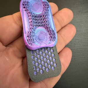 May include: A 3D-printed, multi-colored fidget toy. The top section features two circular indentations and a honeycomb pattern, transitioning from blue to purple. The bottom section is gray with a grid of small, rounded holes. The toy is held in a hand.