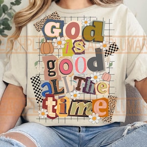 Op de afbeelding: Crèmekleurig T-shirt met de tekst "God is Good All the Time" in collage-stijl. Het ontwerp bevat madeliefjes, pompoenen en geblokte vlag-elementen. De tekst is in verschillende kleuren en lettertypen.