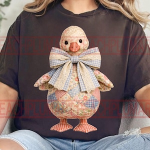 Könnte beinhalten: Eine handgefertigte Entenpuppe mit einem Patchwork-Körper in Rot-, Blau- und Blumenmustertönen. Die Ente hat eine große Schleife mit Spitzentrimm und einem Vichy-Muster. Die Puppe hat einen strukturierten, gemusterten Kopf und Füße.