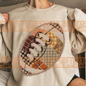 May include: A beige sweatshirt featuring a football design. The football is made of patchwork fabric in shades of brown, orange, and yellow, with a stitched outline. The design is centered on the front of the sweatshirt, offering a cozy, casual look.