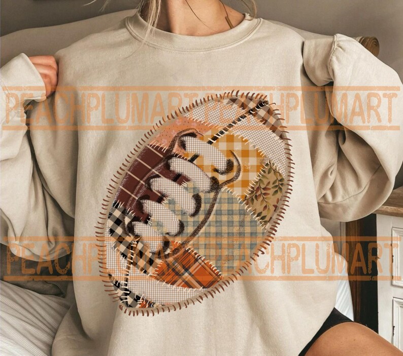 May include: A beige sweatshirt featuring a football design. The football is made of patchwork fabric in shades of brown, orange, and yellow, with a stitched outline. The design is centered on the front of the sweatshirt, offering a cozy, casual look.