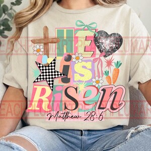 May include: Cream-colored t-shirt with a colorful graphic that reads "He is Risen" with a cross, star, heart, flowers, and carrots. The text "Matthew 28:6" is below the main text. The design has a pastel color scheme.