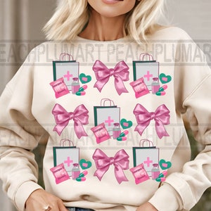 May include: A cream-colored sweatshirt with a pink and green pattern of shopping bags, bows, and medicine bottles. The text "Emergen-C" is visible on the bags.