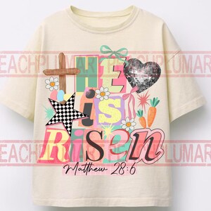 May include: Cream-colored t-shirt with a colorful graphic design. The design includes the words "He is Risen" in various colors, a cross, a star, a heart, carrots, and daisies. The text "Matthew 28:6" is also included.
