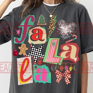 Puede incluir: Camiseta gris oscuro con un gráfico colorido "Fa La La". El diseño incluye un árbol de Navidad, un corazón de bola de discoteca, un hombre de jengibre, una estrella a cuadros y un lazo. La camiseta tiene cuello redondo.