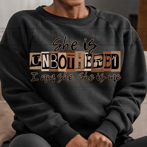 May include: Black sweatshirt with the text "She is Unbothered I am she. She is me" in brown letters. The word "Unbothered" is in a blocky font and each letter is a different shade of brown.