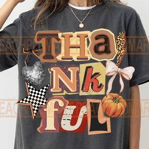 May include: Dark gray t-shirt with a colorful graphic that says "Thankful." The design includes a disco ball heart, a checkered star, a pumpkin, and a bow. The letters are in various colors and fonts.