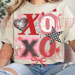 May include: A beige t-shirt with a Valentine's Day design. The design includes the letters "XO" in red and black, a disco ball heart, a checkered star, hearts, bows, and an arrow. The shirt is a casual, everyday wear item.