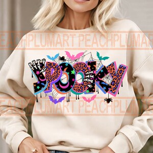 Puede incluir: Una sudadera de color crema con un dise&ntilde;o gr&aacute;fico colorido que dice "Spooky" en una fuente que gotea. El gr&aacute;fico incluye murci&eacute;lagos, ara&ntilde;as, estrellas y un estampado de leopardo.