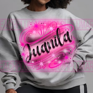 Puede incluir: Una sudadera gris con un dise&ntilde;o aerografiado rosa y negro de un coraz&oacute;n con el nombre Juanita en su interior. El coraz&oacute;n tiene un dise&ntilde;o de espiral y brillos.