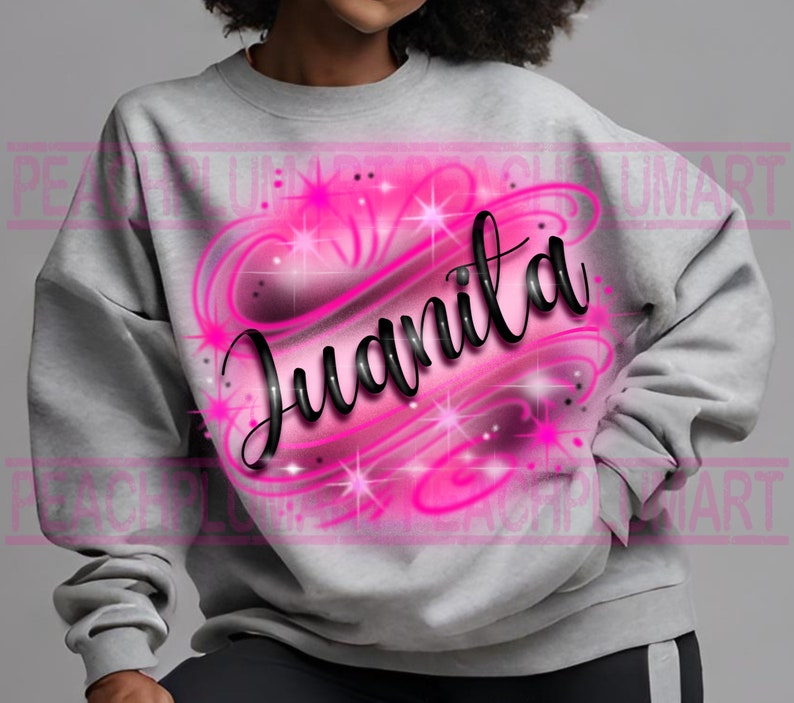 Puede incluir: Una sudadera gris con un dise&ntilde;o aerografiado rosa y negro de un coraz&oacute;n con el nombre Juanita en su interior. El coraz&oacute;n tiene un dise&ntilde;o de espiral y brillos.