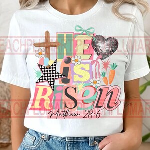 May include: White t-shirt with a colorful graphic design. The design includes the words "He is Risen" in various colors, a wooden cross, a checkered star, a sequined heart, daisies, and carrots. The text "Matthew 28:6" is also included.