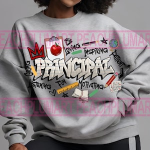 K&ouml;nnte beinhalten: Grauer Pullover mit einem grafischen Design des Wortes "Principal" im Graffiti-Stil. Das Design beinhaltet einen roten Apfel, eine Krone, ein Lineal, ein Notizbuch, einen Bleistift und andere schulbezogene Gegenst&auml;nde. Die W&ouml;rter "Caring, Listening, Fun, Motivating, Loving, Inspiring, Patient" sind ebenfalls im Design enthalten.