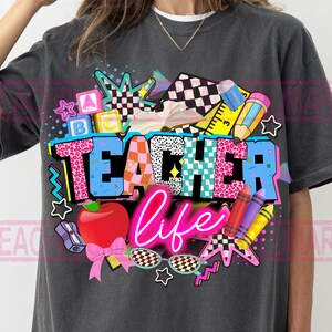 May include: Dark gray t-shirt with a colorful graphic design. The design features the words "TEACHER life" in a vibrant, multi-colored font, along with school-themed illustrations such as an apple, pencils, and stars.