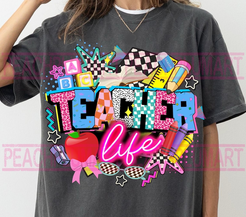 May include: Dark gray t-shirt with a colorful graphic design. The design features the words "TEACHER life" in a vibrant, multi-colored font, along with school-themed illustrations such as an apple, pencils, and stars.