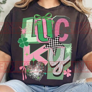 May include: Dark brown t-shirt with a graphic design featuring the word "LUCKY" in green, pink, and gray. The design includes a four-leaf clover, a disco ball heart, a checkered star, and other decorative elements. The shirt is a casual wear item.