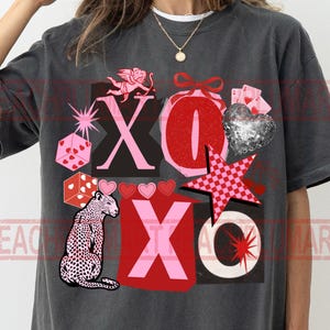 May include: Dark gray t-shirt with a graphic design featuring the letters "XO", a leopard, dice, hearts, a disco ball heart, and a star. The design incorporates pink, red, and white elements.