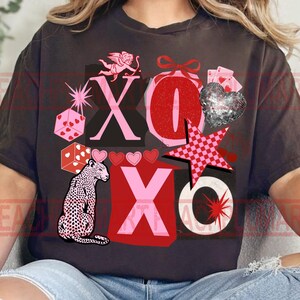 May include: Dark gray t-shirt with a Valentine's Day graphic. The design includes the letters "XO", a leopard, hearts, dice, a star, and a cupid. The colors are red, pink, and black.