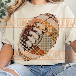 May include: Cream-colored t-shirt featuring a football design. The football is a patchwork of various patterns, including plaid and floral prints, in shades of brown, orange, and beige. The design is stitched with a dark thread.