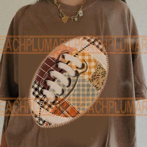 May include: Brown t-shirt featuring a patchwork football design. The football is made of various patterned fabrics in shades of brown, orange, and cream. The shirt has short sleeves and a relaxed fit, suitable for casual wear.