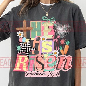 May include: Dark gray t-shirt with a colorful graphic design. The design includes a wooden cross, a disco ball heart, and the words "He Has Risen" with the Bible verse Matthew 28:6.